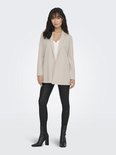 Vrouw met lange, golvende donkere haren, staand in een lange, beige blazer, witte top en zwarte broek.