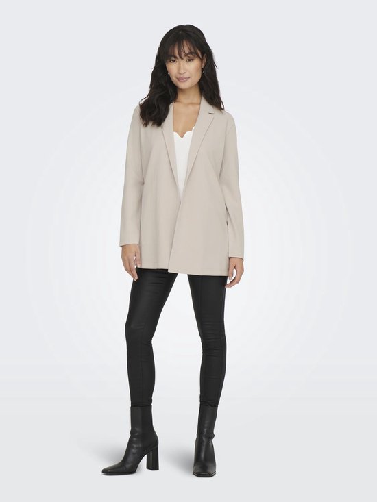 Vrouw met lange, golvende donkere haren, staand in een lange, beige blazer, witte top en zwarte broek.