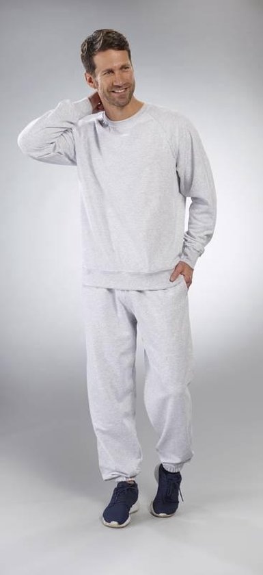 Man draagt een lichtgrijze trui met ronde hals en een lichtgrijze joggingbroek. Hand in de zak.