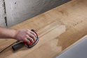 Hand schuurt onbehandeld hout met een schuurmachine, waarbij de houtnerf zichtbaar wordt.