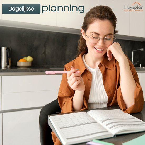 Vrouw met bril en oranje blouse kijkt glimlachend naar een open budgetplanner, terwijl ze een pen vasthoudt. Ze zit aan een tafel in een moderne keuken.