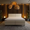 Een beige boxspring met een gestoffeerd hoofdbord met vier vakken, een matras met honingraatpatroon en twee nachtkastjes met lampen.