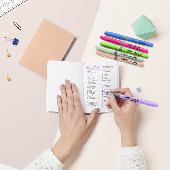 Handen die met een paarse BIC markeerstift schrijven in een klein notitieboekje, naast andere pastelkleurige markeerstiften, een mapje en kantoorbenodigdheden.