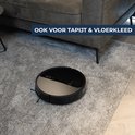 Zwarte robotstofzuiger op dik grijs vloerkleed naast grijze bank.