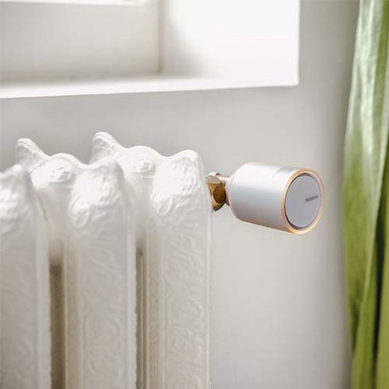 Netatmo slimme thermostaatknop, zilverkleurig met oranje accent, op een oude crèmekleurige radiator.