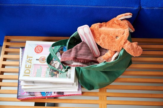 Een lichtbeige Mepal babylepelsset met twee lepels in een houder, liggend in een groene tas naast een stapel tijdschriften en een knuffel.