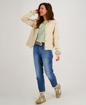 Vrouw met bruin haar, draagt een beige bomberjack, groen-wit gestreepte top, blauwe jeans en bruine riem.