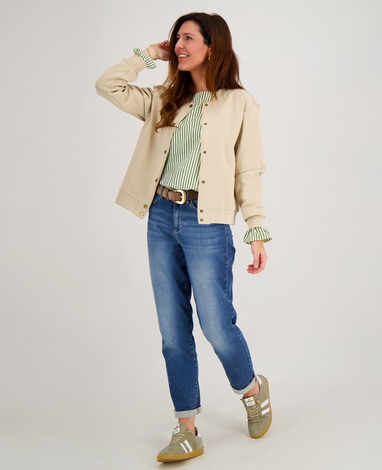 Vrouw met bruin haar, draagt een beige bomberjack, groen-wit gestreepte top, blauwe jeans en bruine riem.