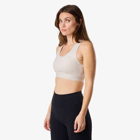 Model van de zijkant, draagt een lichte sportbeha met racerback en zwarte legging met logo. Ze kijkt naar de camera.
