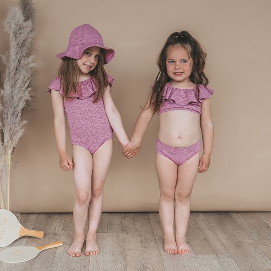 Twee meisjes hand in hand, één in roze badpak met broderie, ander in roze bikini met broderie.