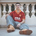 Man met kort donker haar en snor, zittend in een AS Roma retro voetbalshirt in wijnrood met oranje kraag.