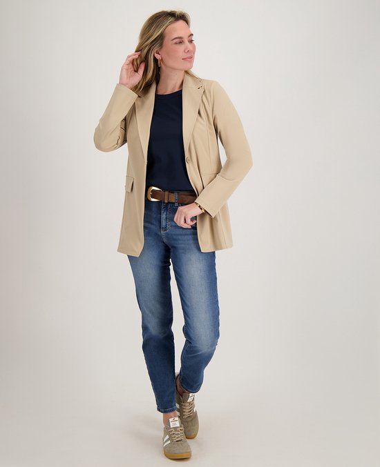 Model draagt een beige blazer, donkerblauw T-shirt, spijkerbroek en sneakers.