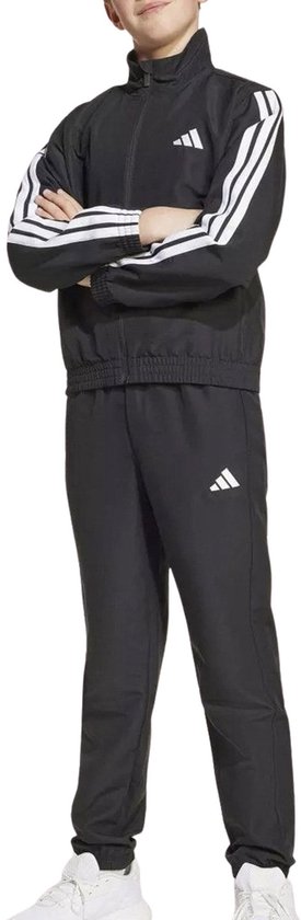 Jongen met armen over elkaar in zwart Adidas trainingspak met witte strepen op de mouwen en logo op jas en broek.