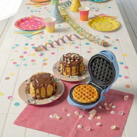 Blauw wafelijzer op een roze onderlegger, met gestapelde wafels gegarneerd met chocolade en snoepjes.