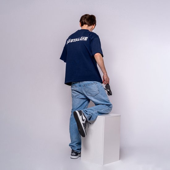 Jonge man met bruin haar, staand met zijn rug naar de camera, leunend op een witte kubus. Hij draagt een donkerblauw t-shirt met opdruk 'NOSTALGIE' en een spijkerbroek.