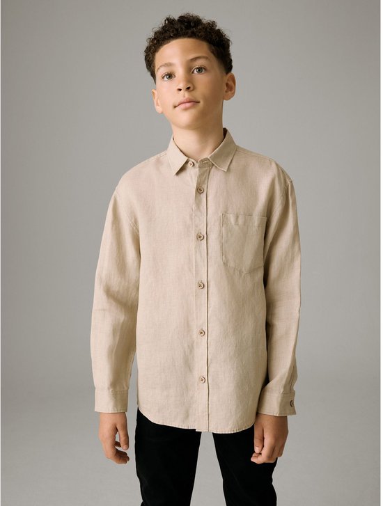 Jongen in een beige linnen overhemd met lange mouwen en een borstzakje, gedragen met een zwarte broek.