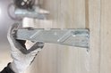 Een hand met een grijze werkhandschoen houdt een metalen montagebeugel vast tegen een betonnen muur.