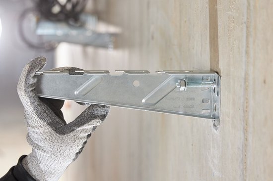 Een hand met een grijze werkhandschoen houdt een metalen montagebeugel vast tegen een betonnen muur.