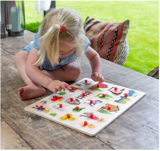 Blond meisje met een blauw jurkje en strik in haar haar, speelt met een houten boerderijpuzzel op een tafel.