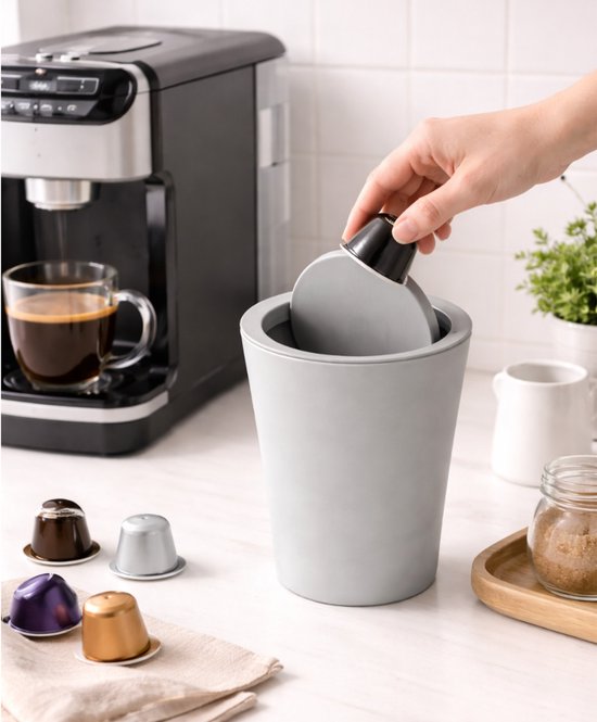 Grijze miniprullenbak met deksel, naast een koffiezetapparaat. Een hand deponeert een koffiecup.
