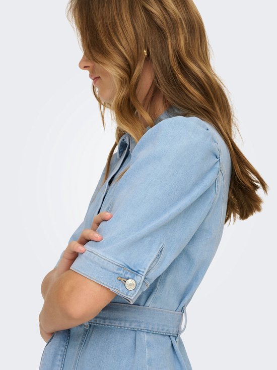 Model draagt een lichtblauwe spijkerjurk met korte pofmouwtjes met knoopjes en een ceintuur.
