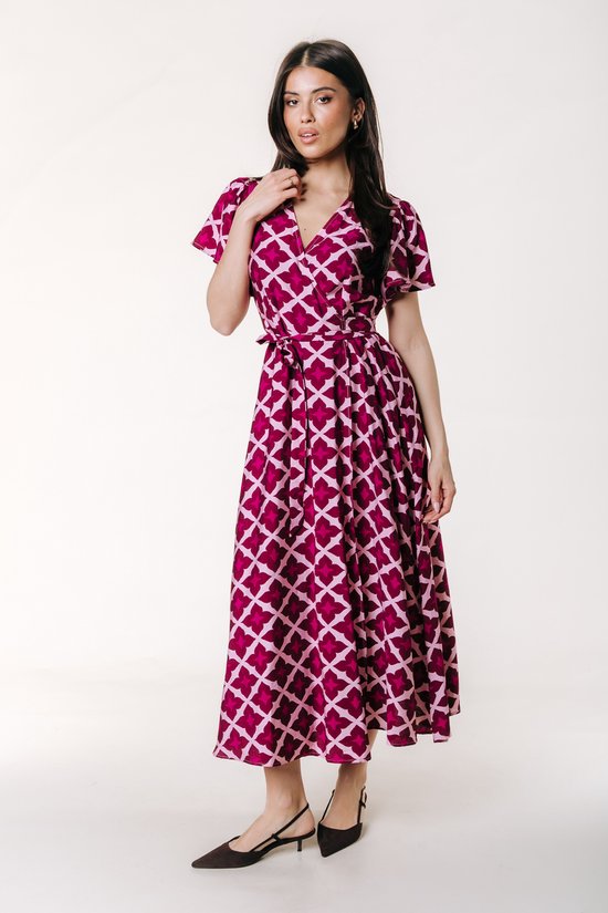 Model in een lange wikkeljurk met korte pofmouwen en een geometrisch patroon in roze en paars. De jurk heeft een strikceintuur.