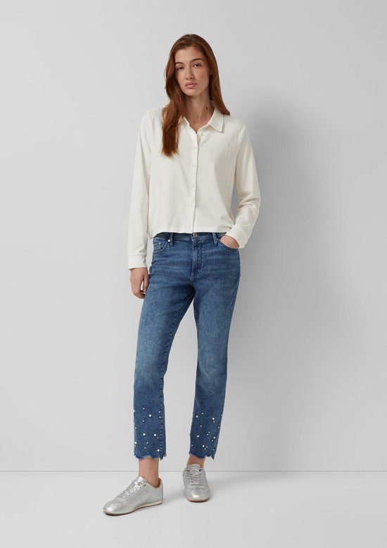 Model staat in een witte blouse en zilveren sneakers. Ze draagt een blauwe jeans met pareltjes en rafels aan de onderkant.