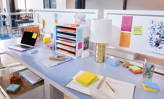 Een bureau vol met kantoorartikelen, waaronder Post-it notities, een laptop en een lamp.