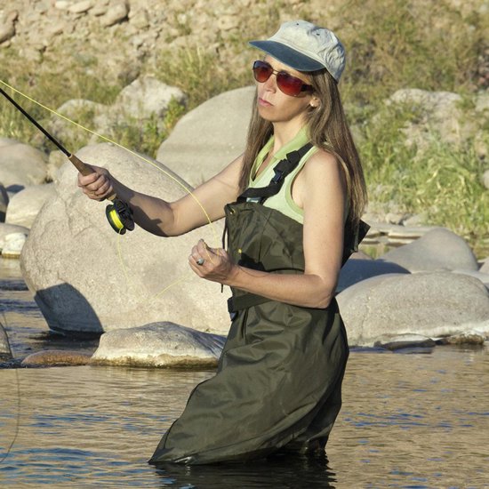 Vrouw in olijfgroene waadbroek, vissend in een ondiepe rivier, met grote stenen op de achtergrond.