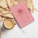 Roze A5 budget planner naast een kop koffie op een onderzetter, met een sjaal op de achtergrond.