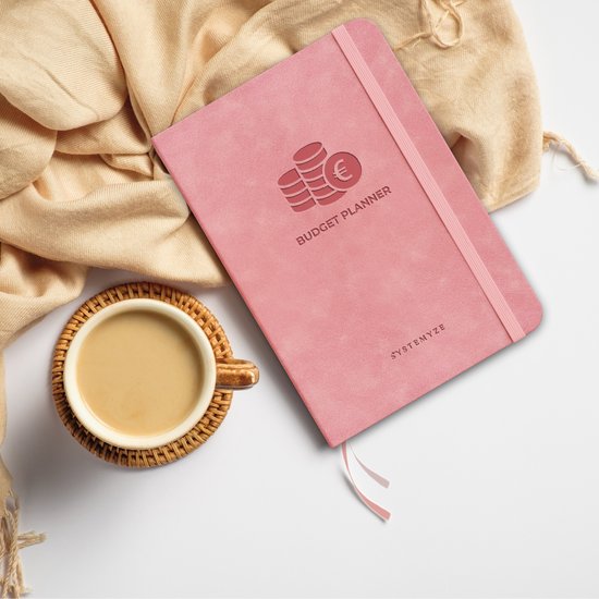 Roze A5 budget planner naast een kop koffie op een onderzetter, met een sjaal op de achtergrond.