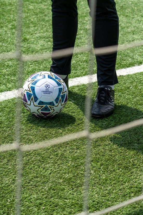 Voetbal met UEFA Champions League logo en sterrenpatroon, op groen gras. Een persoon in zwarte broek en schoenen staat ernaast.