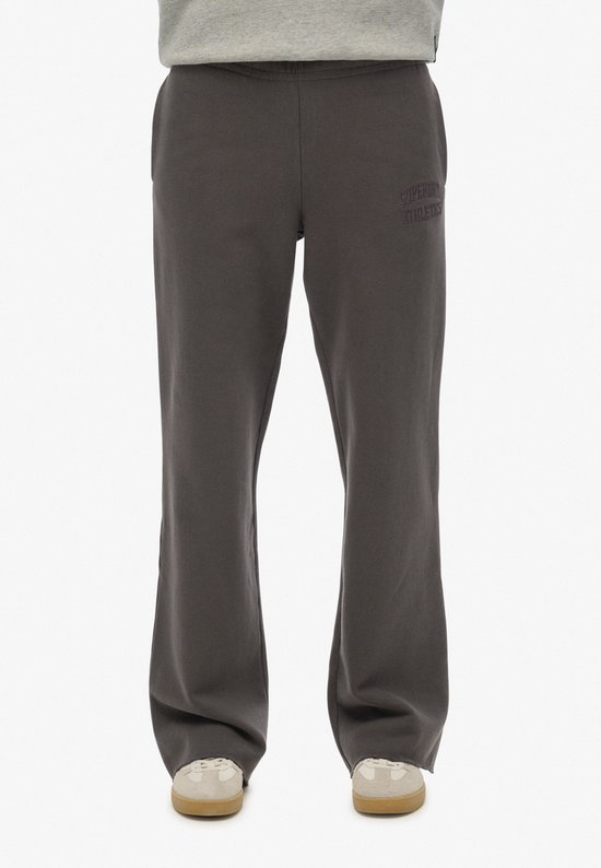 Een model draagt een donkergrijze joggingbroek met wijde pijpen en geborduurde tekst 'Superdry Athletics'.
