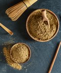 Houten kom gevuld met matcha poeder, met een houten schepje, een matcha klopper en een bamboe lepel ernaast.