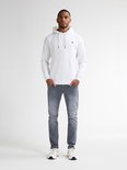 Man van voren gezien, draagt een witte hoodie met ronde hals en grijze slim fit jeans.