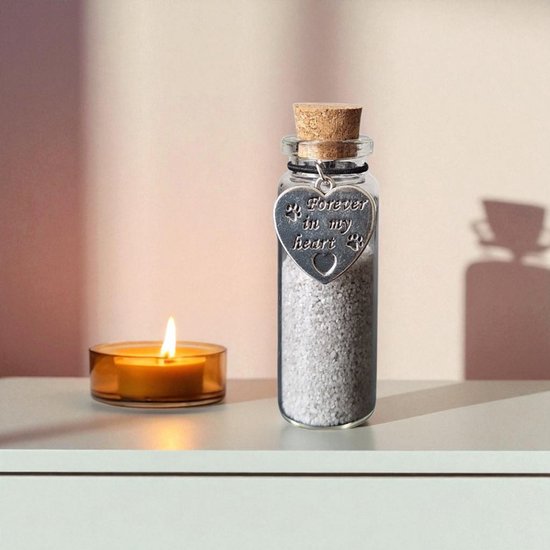 Glazen flesje met kurk, gevuld met lichtgrijs zand en een zilveren hartvormige bedel met 'Forever in my heart' en pootafdrukken, naast een brandende kaars.