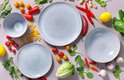 Stoneware borden en kommen in lichtblauw met gespikkelde afwerking, omringd door verse ingrediënten zoals groenten en pasta.