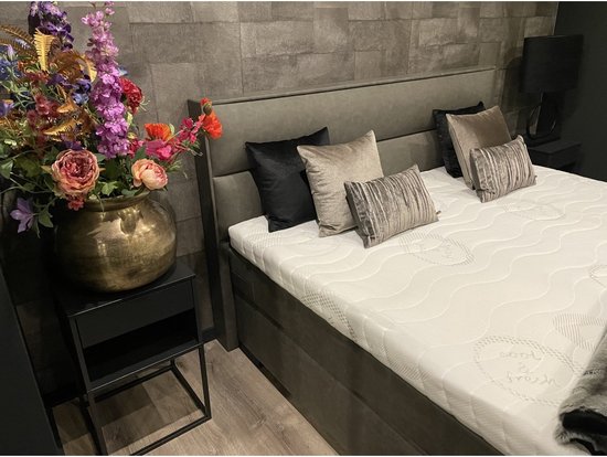 Een boxspring met een grijs hoofdbord en bijpassende matrassen. De afbeelding toont een vaas met bloemen op een nachtkastje naast het bed.