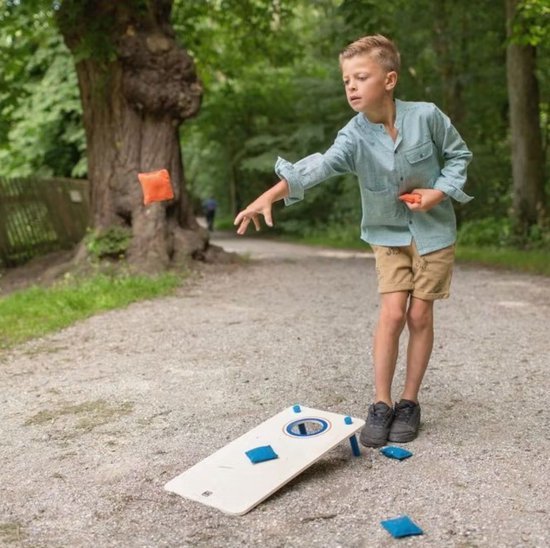 Jongen gooit oranje beanbag naar houten cornhole board met blauwe beanbags, in bosrijke omgeving.