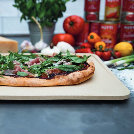 Pizza met ham, rucola, parmezaan op een beige pizzasteen in een keukenomgeving.