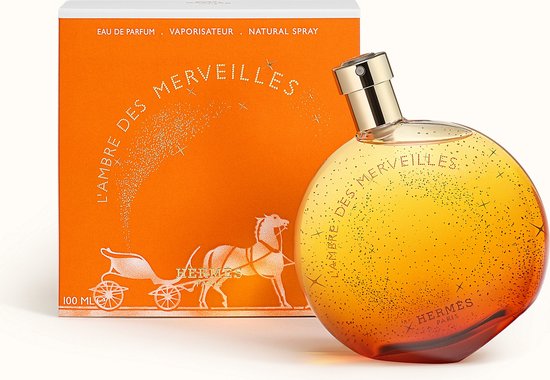 Oranje parfumfles met gouden dop, met sprankelende gouden deeltjes en L'Ambre des Merveilles tekst. Naast een oranje doosje met karos en paard.
