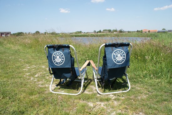 Twee donkerblauwe campingstoelen met witte logo's, in een graslandschap met water op de achtergrond.