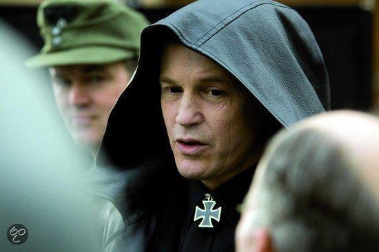 Man met grijze kap en kruisvormig medaillon, in de verte een man in militair uniform.