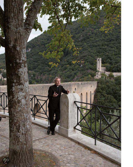 Antoine Bodar, in een zwarte jas, staat op een balkon met een stenen balustrade, met een oude brug en een kasteel op de heuvels op de achtergrond.