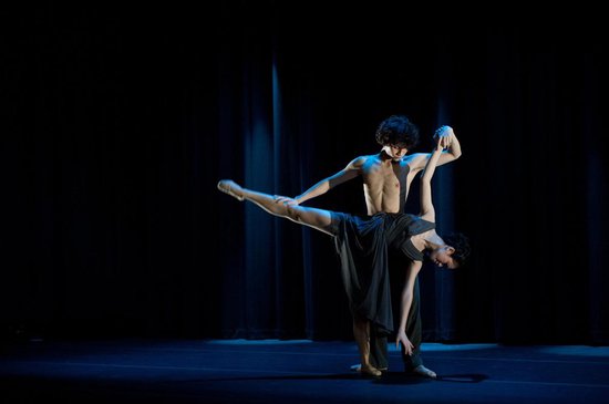 Een man en vrouw dansen ballet in zwarte kleding op een donker podium. De man houdt de vrouw vast terwijl ze op één been staat.