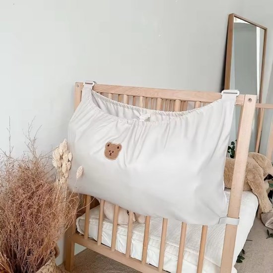 Lichtkleurige hangende organizer met teddybeerlogo, bevestigd aan een houten ledikant.