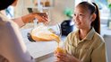 Hand schenkt oranje smoothie uit blenderkan in glas, meisje kijkt toe.