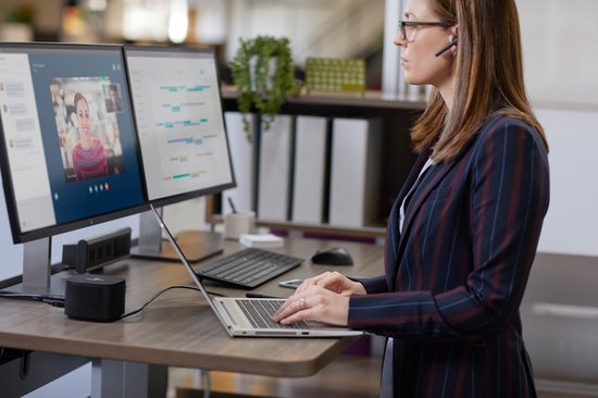 Zakenvrouw in gestreept jasje met headset staat achter een bureau met twee monitoren.