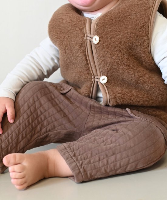 Baby met bruine teddy vest, witte lange mouwen en bruine broek.