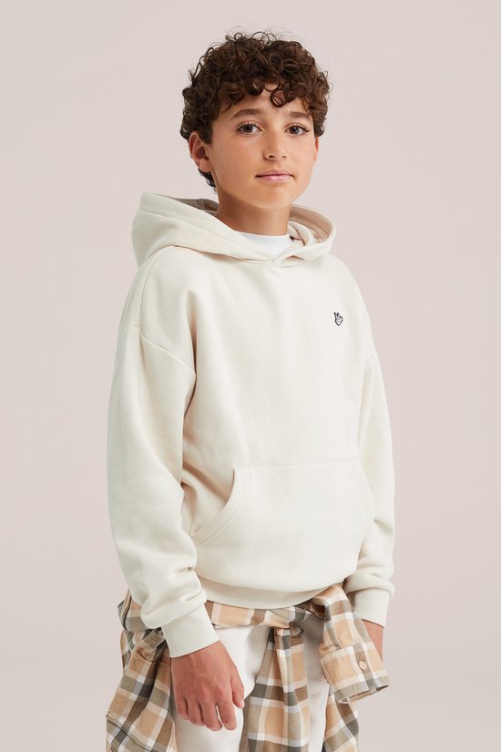 Jongen draagt een beige hoodie met handborduurwerk, gecombineerd met een geruit overhemd rond de middel.